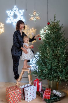 A mother with a 2-year-old daughter decorates the Christmas tree. Mom in a black suit, a girl in a white dress, her daughter hangs a red ball on the Christmas tree. Merry Christmas and New Year concept.