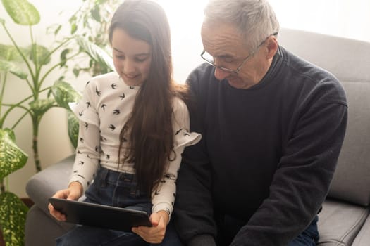 Happy family senior grandfather and girl watching cartoon on tablet on weekend day at home together.