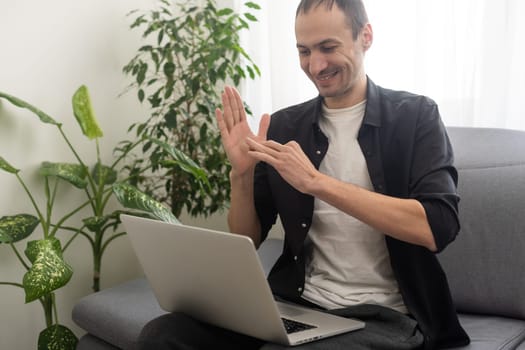 Happy smiling deaf young caucasian man uses sign language while video call using laptop while sitting at home, virtual communication concept