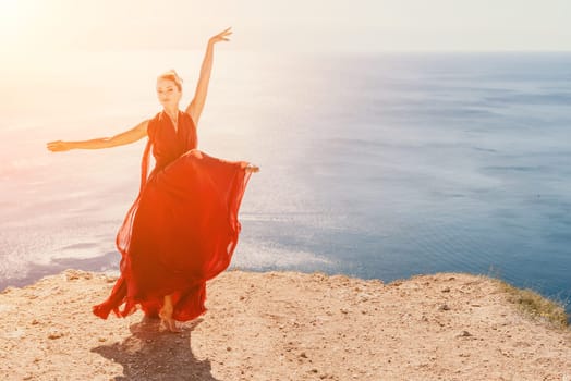 Side view a Young beautiful sensual woman in a red long dress posing on a rock high above the sea during sunrise. Girl on the nature on blue sky background. Fashion photo.