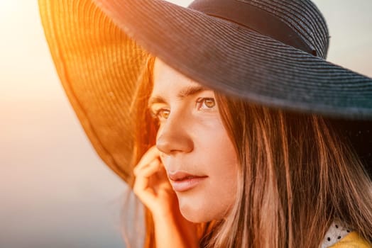 Portrait of happy young woman wearing summer black hat with large brim at beach on sunset. Closeup face of attractive girl with black straw hat. Happy young woman smiling and looking at camera at sea