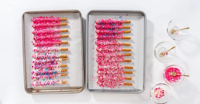 Flat lay. Drizzling melted chocolate over chocolate-dipped pretzels rods and decorating with sprinkles to make chocolate-covered pretzel rods for Valentine's Day.