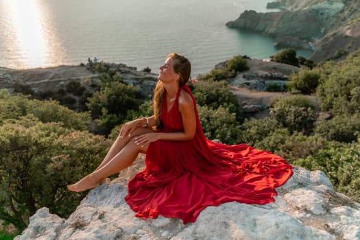 Woman sunset sea red dress, side view a happy beautiful sensual woman in a red long dress posing on a rock high above the sea on sunset