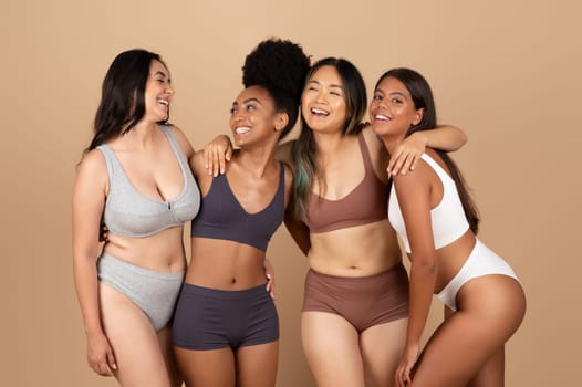 Diverse women showcase their natural beauty in understated underwear, sharing a moment of joy and unity, against beige studio backdrop