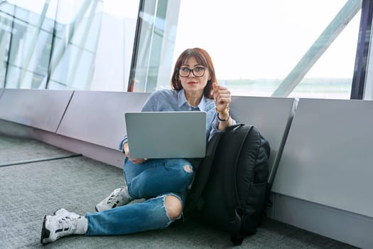 Middle aged woman with backpack waiting for airplane flight in airport terminal, using laptop, looking at camera. Travel, business trip, passenger air transport concept