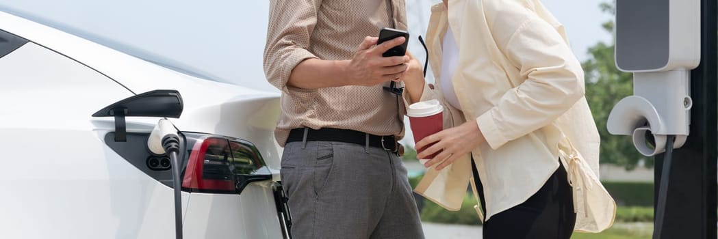 Young couple with coffee cup, recharge electric car's battery from EV charging station in green city park. Sustainable and eco friendly EV car with urban and shopping lifestyle. Panorama Expedient