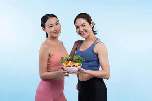 Two young sporty Asian women in sportswear holding salad bowl fill with fruit and vegetable. Natural youthful and fit body lifestyle people with balance nutrition on isolated background. Vigorous