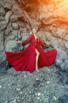 Red dress rocks woman. A blonde with flowing hair in a long flowing red dress stands near a rock of volcanic origin. Travel concept, photo session at sea.