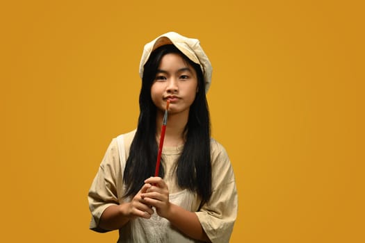 Cheerful teenage Asian girl holding palette and paint brushes on yellow background. Art, creativity and education concept.