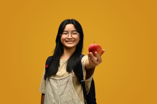 Happy schoolgirl wearing glasses holding an apple on yellow background. Educational, learning and back to school concept.