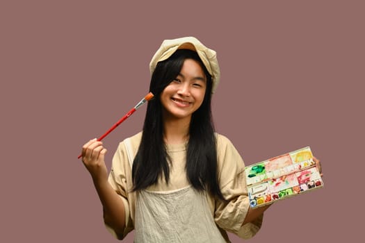 Cute teenage girl holding palette and paint brushes on pink background. Art, creativity and education concept.