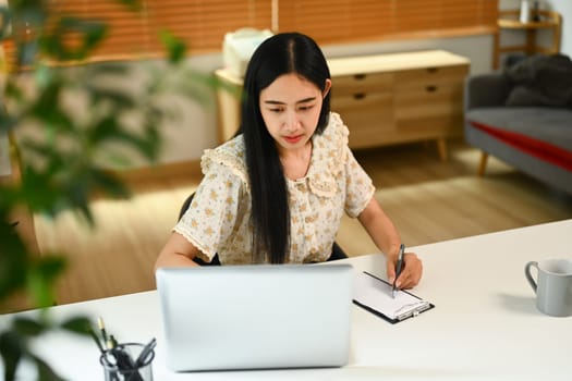 Young woman freelancer looking at laptop screen and writing information on paper..