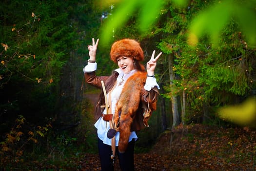 Girl in a leather jacket, a big red fox fur hat and with the skin of a fox killed on the hunt in the forest in autumn. A female model poses as fabulous royal huntress on nature hunt at photo shoot