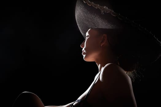 Silhouette of a beautiful young girl in black underwear and a Mexican hat on dark background. Exotic model posing naked in the studio. Body portrait of sexy girl in usial photoshoot