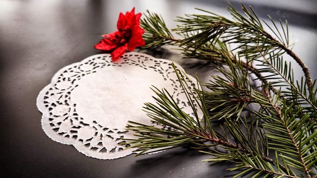 Black frame, white round napkin, green pine branch, red flower on background with reflection and sparkle of light. Abstract location for photo shoot. Cchristmas table decor. Concept of winter holiday