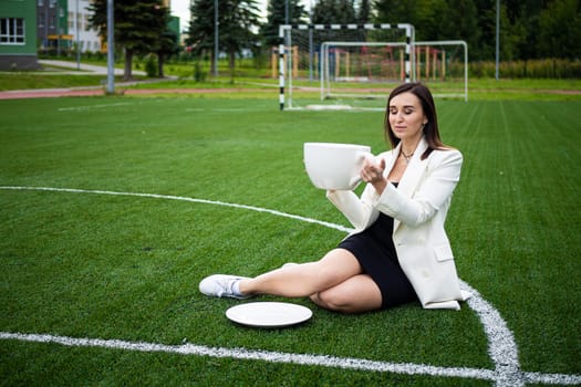 A business woman with a large cup, sitting on a green lawn in the park. The concept of an office worker on a picnic.