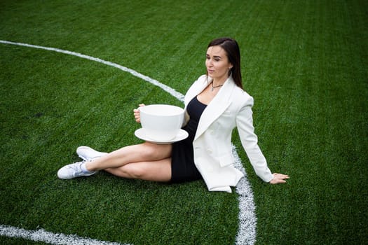 A business woman with a large cup, sitting on a green lawn in the park. The concept of an office worker on a picnic.