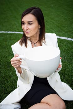 A business woman with a large cup, sitting on a green lawn in the park. The concept of an office worker on a picnic.