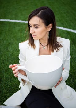 A business woman with a large cup, sitting on a green lawn in the park. The concept of an office worker on a picnic.