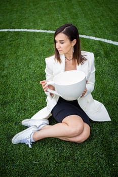 A business woman with a large cup, sitting on a green lawn in the park. The concept of an office worker on a picnic.