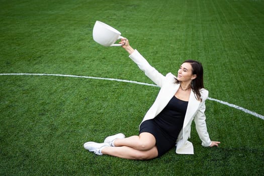 A business woman with a large cup, sitting on a green lawn in the park. The concept of an office worker on a picnic.