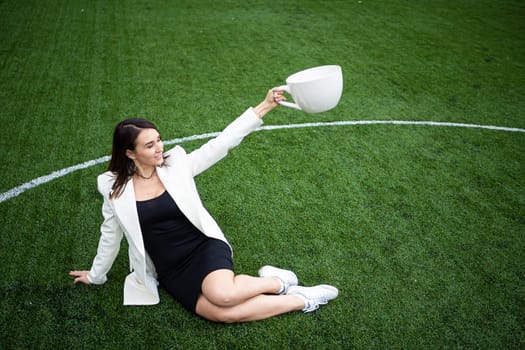 A business woman with a large cup, sitting on a green lawn in the park. The concept of an office worker on a picnic.