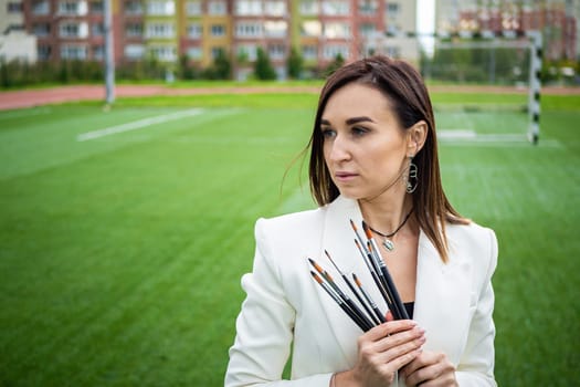 Brunette, female artist paints in nature. She has paint brushes in her hands.