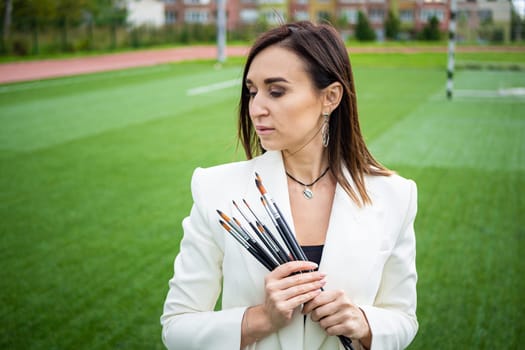 Brunette, female artist paints in nature. She has paint brushes in her hands.
