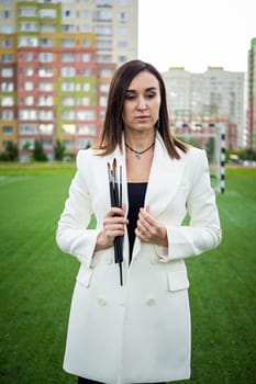 Brunette, female artist paints in nature. She has paint brushes in her hands.