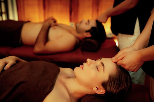 Couple customer enjoying relaxing anti-stress head massage and pampering facial beauty skin recreation leisure in warm candle lighting ambient salon spa in luxury resort or hotel. Quiescent