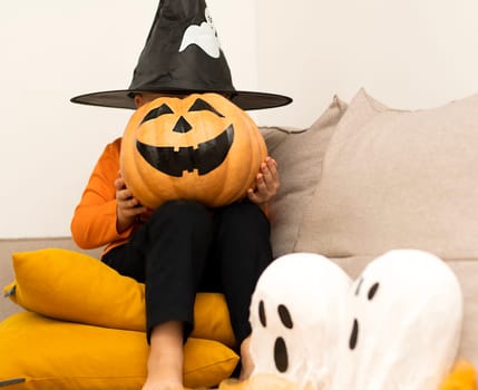 Halloween. Concept. A boy in an orange sweater and a haunted wizard's hat, with a pumpkin instead of a face, sits cheerfully in a home interior. Close-up