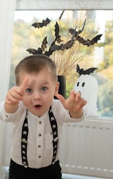 Halloween concept. A little boy, with emotional facial expressions, in a white shirt and black suspenders with images of skulls against the background of a window with a vase of dry branches and black paper bats. Close-up