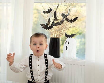 Halloween concept. A little boy, with emotional facial expressions, in a white shirt and black suspenders with images of skulls against the background of a window with a vase of dry branches and black paper bats. Close-up