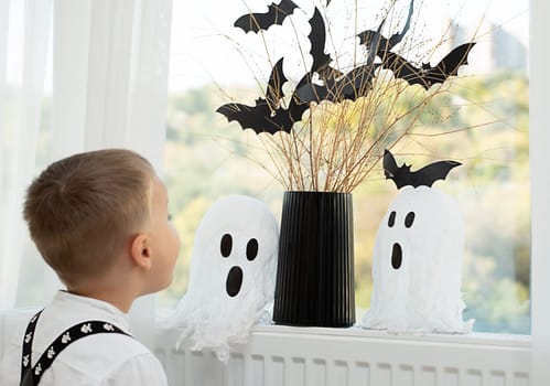 Halloween concept. A little boy, with emotional facial expressions, in a white shirt and black suspenders with images of skulls against the background of a window with a vase of dry branches and black paper bats. Close-up