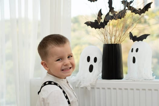 Halloween concept. A little boy, with emotional facial expressions, in a white shirt and black suspenders with images of skulls against the background of a window with a vase of dry branches and black paper bats. Close-up