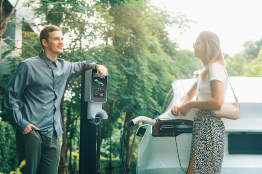 Young couple travel with EV electric car charging in green sustainable city outdoor garden in summer shows urban sustainability lifestyle by green clean rechargeable energy of electric vehicle innards