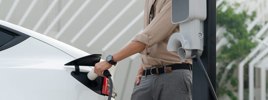 Young man recharge electric car's battery from charging station in city commercial parking lot. Rechargeable EV car for sustainable environmental friendly urban travel. Panorama Expedient