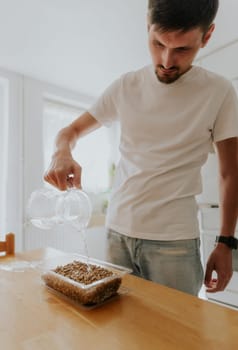 One young Caucasian recognizable man waters wheat seeds with soil into a container with one hand from a glass jug, standing in the kitchen at a wooden table, close-up side view.