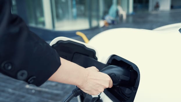 Progressive businesswoman install charger plug from charging station to his electric car before driving around city center. Eco friendly rechargeable car powered by sustainable and clean energy.