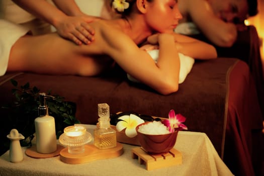 Caucasian couple customer enjoying relaxing anti-stress spa massage and pampering with beauty skin recreation leisure in warm candle lighting ambient salon spa at luxury resort or hotel. Quiescent