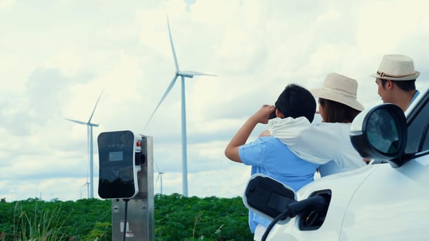 Concept of progressive happy family enjoying their time at wind farm with electric vehicle. Electric vehicle driven by clean renewable energy from wind turbine generator for charging station.