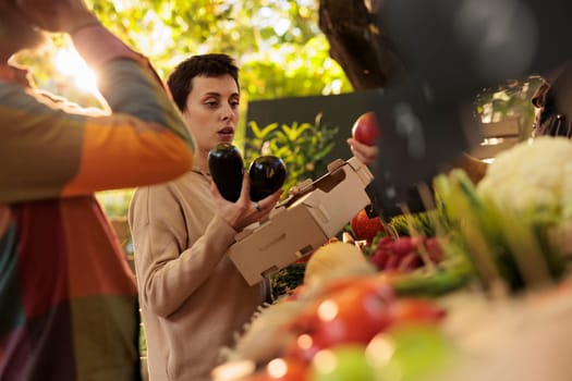 Young woman vegetarian shopping at farmers market. Female customer putting fresh organic seasonal fruits and vegetables in cardboard box, buying healthy locally-grown produce