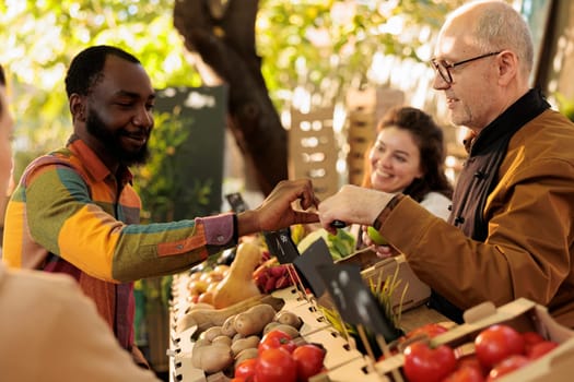 Senior farmer giving apple sample to african american man, client trying out bio organic fruits and vegetables. Old person small business owner selling organic eco products at farm stand.