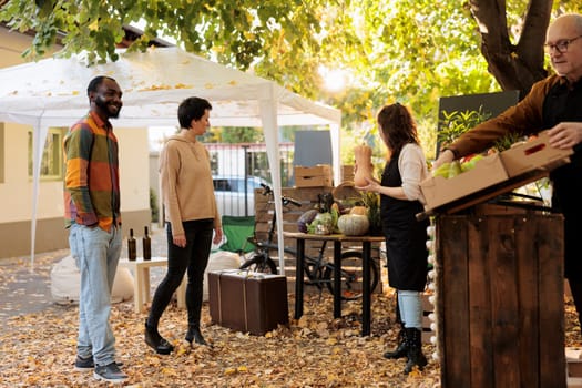 Young multiracial family couple visit farmers market to shop for healthy local food. Vendors standing near organic produce stand greeting first customers, offering locally grown fruits and veggies