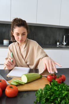 Portrait of woman writing down list of groceries, making notes in recipe, sitting in kitchen near vegetables, preparing dinner menu.