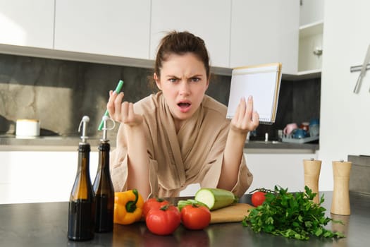 Portrait of woman with angry face, standing near vegetables and looking frustrated, holding notebook, annoyed while cooking meal. Lifestyle and people concept
