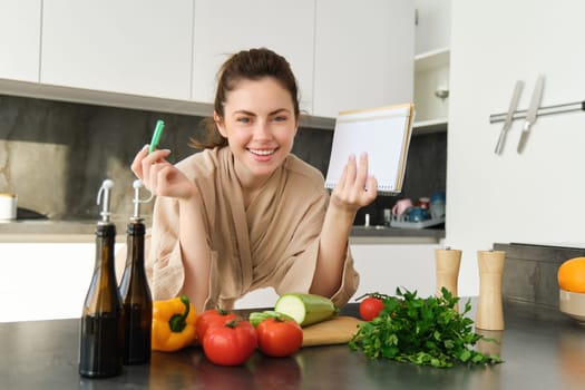 Smiling young woman in bathrobe, cooking in the kitchen, standing near vegetables with notebook, writing down recipe, checking grocery list, cooking fresh and healthy meal.