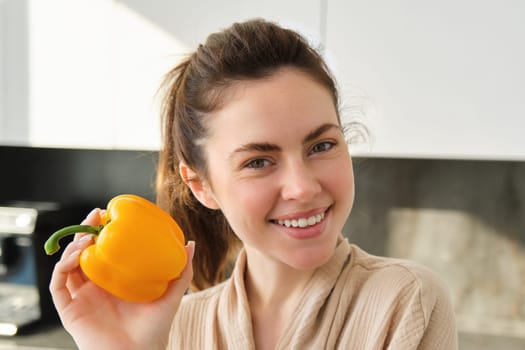 Close up portrait of beautiful woman smiling, showing yellow pepper, vegetarian girl making dinner, preparing meal with vegetables, posing in kitchen.