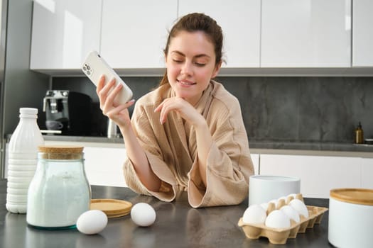 Attractive young cheerful girl baking at the kitchen, making dough, holding recipe book, having ideas.