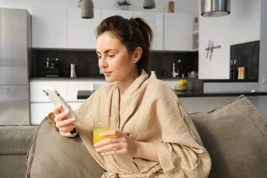 Lifestyle and morning concept. Beautiful woman sitting on sofa and resting, reading news on smartphone, drinking orange juice at home.